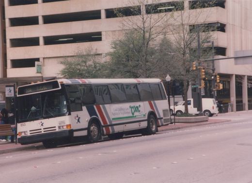A white Fort Worth Transit bus with red and blue stripes, number 853, is stopped on a city street. A green and white advertisement is on its side, and a multi-story parking garage is visible in the background.