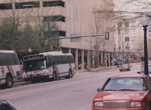 A white Fort Worth Transit bus with red and blue stripes, number 506, travels down a city street with a red car in the foreground and a large building in the background.