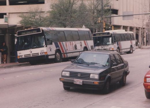 Two white buses with red and blue stripes, one numbered 824, are on a city street with a dark gray Volkswagen Jetta in the foreground and a few people waiting at a bus stop.