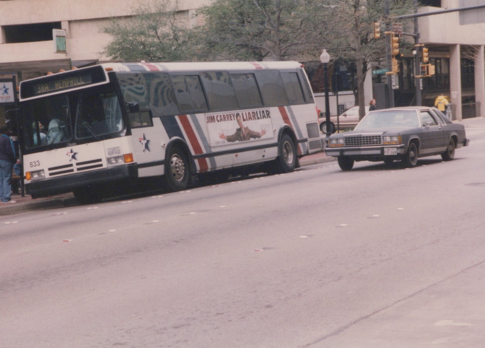 A white city bus with red and blue stripes and a star logo is stopped at a curb on a city street. A large ad for the movie 'Liar Liar' runs along its side. A dark-colored sedan and a few people are nearby.