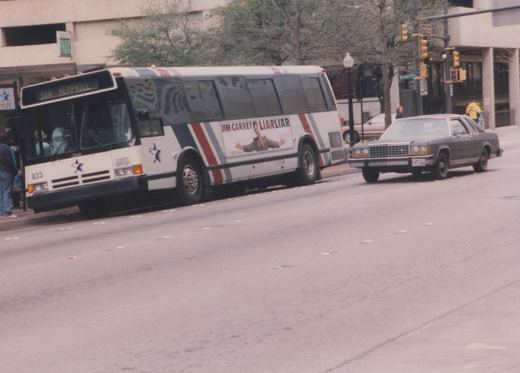 A white city bus with red and blue stripes and a star logo is stopped at a curb on a city street. A large ad for the movie 'Liar Liar' runs along its side. A dark-colored sedan and a few people are nearby.
