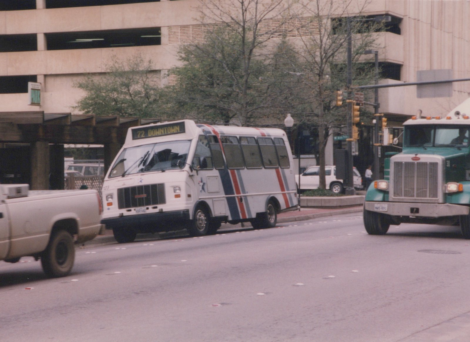 A white bus with blue, red and grey stripes and a star logo is stopped at an intersection in a city. A green semi-truck and other vehicles are nearby, and a parking garage is in the background.