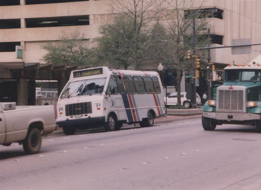 A white bus with blue, red and grey stripes and a star logo is stopped at an intersection in a city. A green semi-truck and other vehicles are nearby, and a parking garage is in the background.