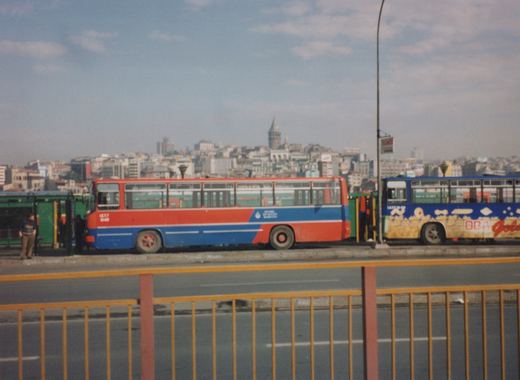 Two colorful motor buses, one red and blue and one blue with advertising, stand on a bridge in Istanbul with a cityscape and a historic tower in the background.