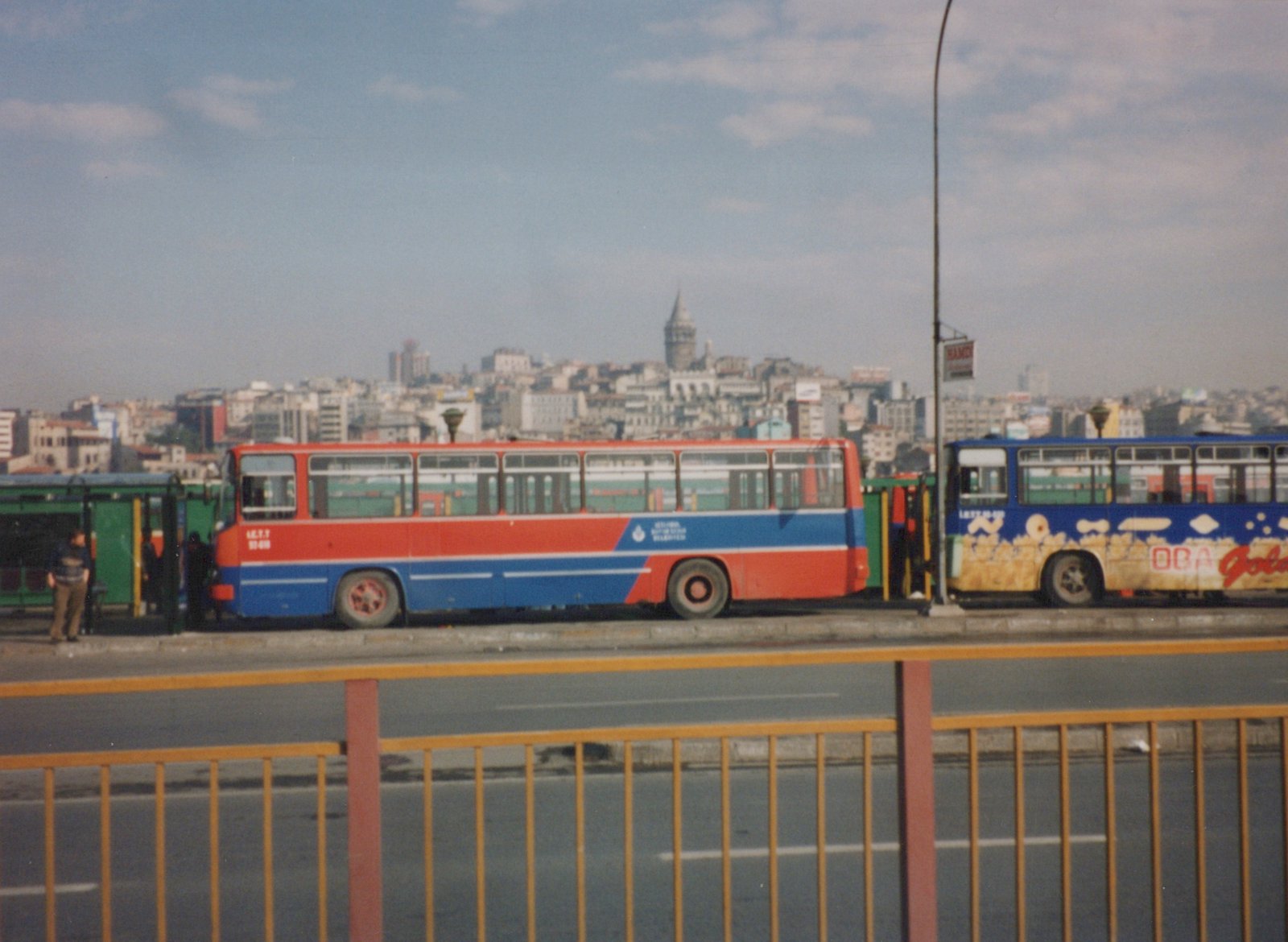 Two colorful motor buses, one red and blue and one blue with advertising, stand on a bridge in Istanbul with a cityscape and a historic tower in the background.
