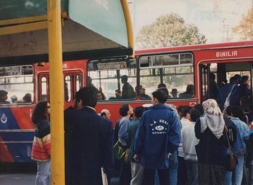 A red and blue bus with the lettering 'BİNİLİR' and route number '29C' is stopped at a curb with passengers boarding. A crowd of people, including a man in a blue jacket and a woman in a headscarf, wait nearby. A yellow pole and some trees are in the background.