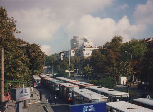 A high-angle view of a street in Istanbul filled with buses, some red and white and others white, with passengers and pedestrians nearby, and a tall building with a 'CONRAD' sign in the background.