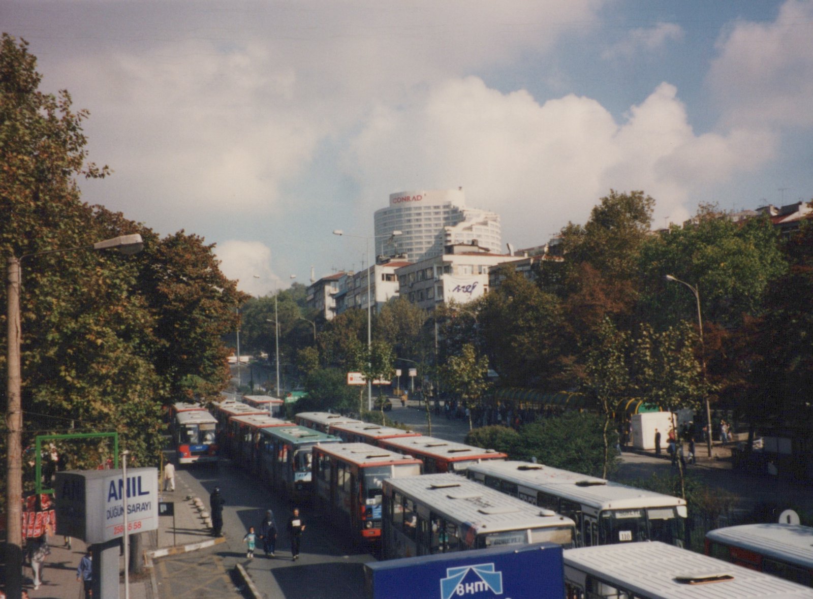 A high-angle view of a street in Istanbul filled with buses, some red and white and others white, with passengers and pedestrians nearby, and a tall building with a 'CONRAD' sign in the background.