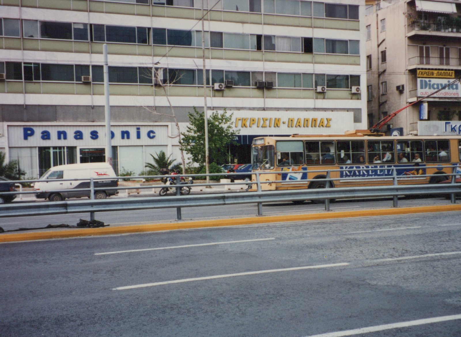A yellow trolleybus with passengers drives down a city street past a Panasonic store and other buildings; a white van and a motorcycle are also on the road.