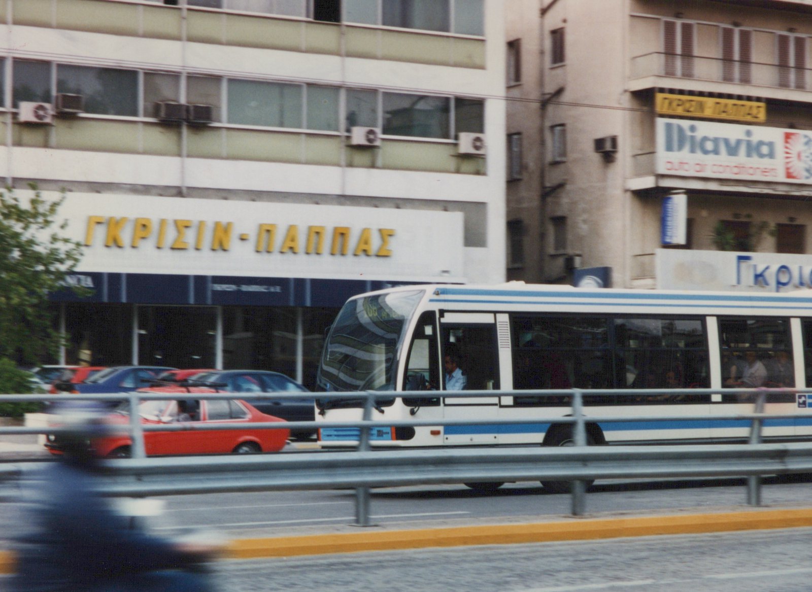 A side view of a white and blue Neoplan bus driving on a city street, with a red car and storefronts visible behind it.