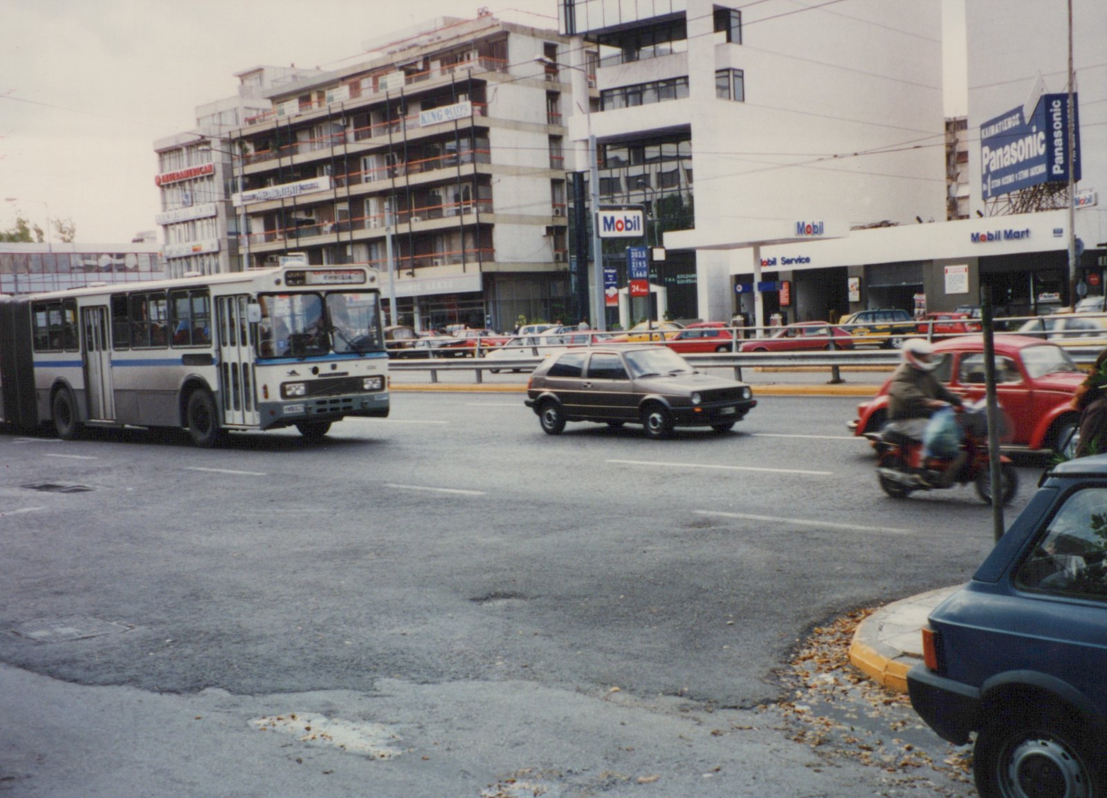 A white and blue articulated bus drives down a street beside a Mobil gas station, with a brown Volkswagen hatchback and a red motorcycle nearby.