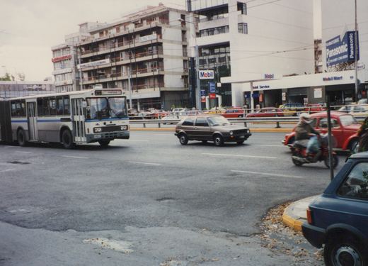 A white and blue articulated bus drives down a street beside a Mobil gas station, with a brown Volkswagen hatchback and a red motorcycle nearby.