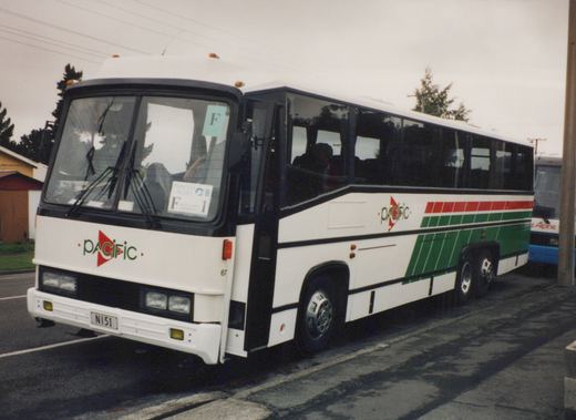 A white Pacific motor bus with green and red stripes, number 67, parked on a wet street on an overcast day, with a blue bus partially visible behind it.