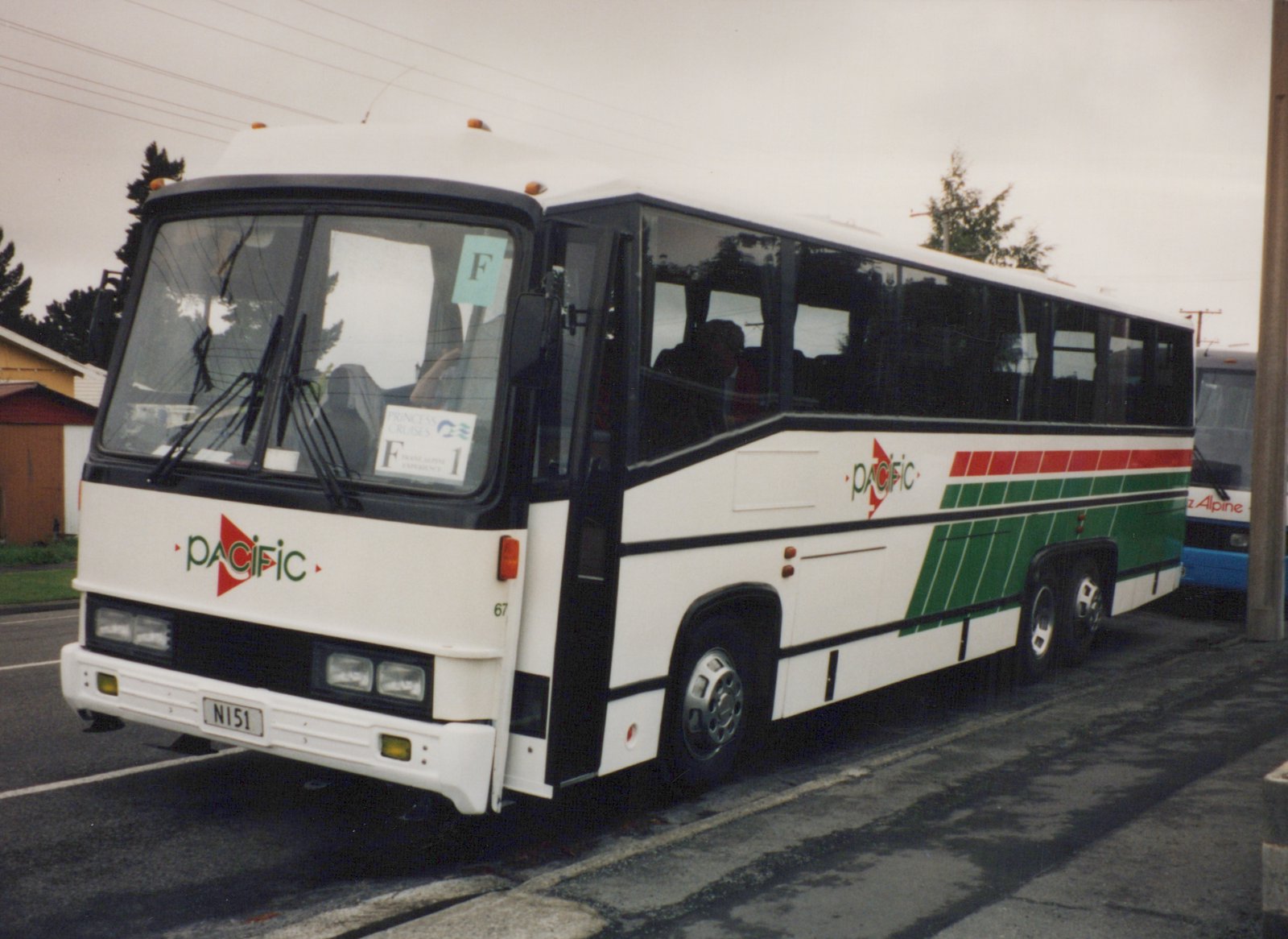 A white Pacific motor bus with green and red stripes, number 67, parked on a wet street on an overcast day, with a blue bus partially visible behind it.