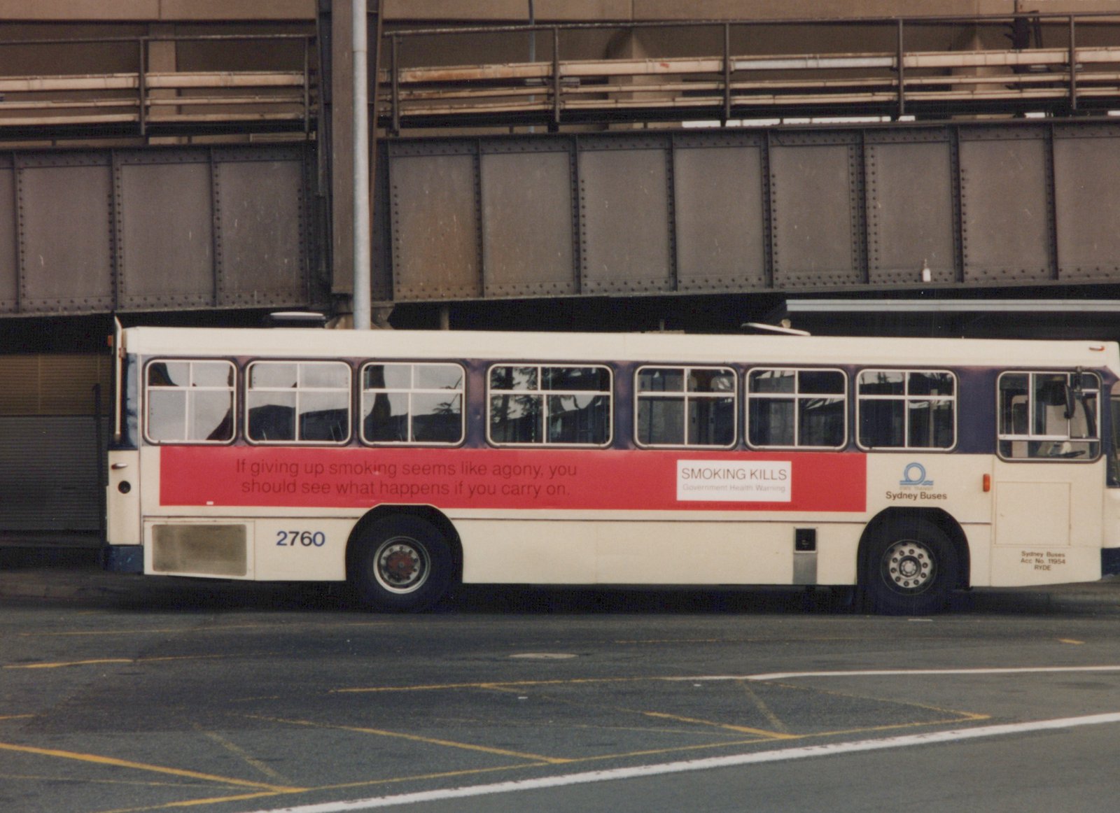 Side view of a white and blue Sydney Buses motor bus with number 2760, parked under a steel railway bridge with a red anti-smoking ad on its side.