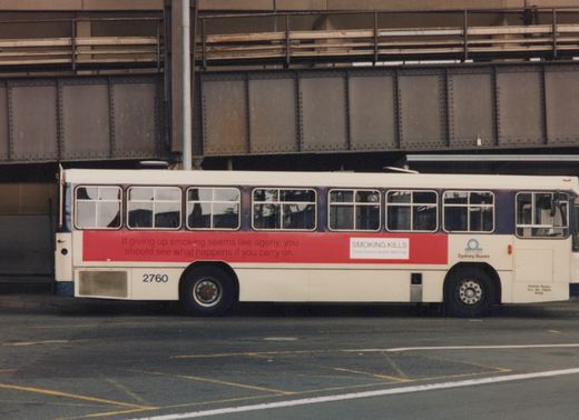 Side view of a white and blue Sydney Buses motor bus with number 2760, parked under a steel railway bridge with a red anti-smoking ad on its side.