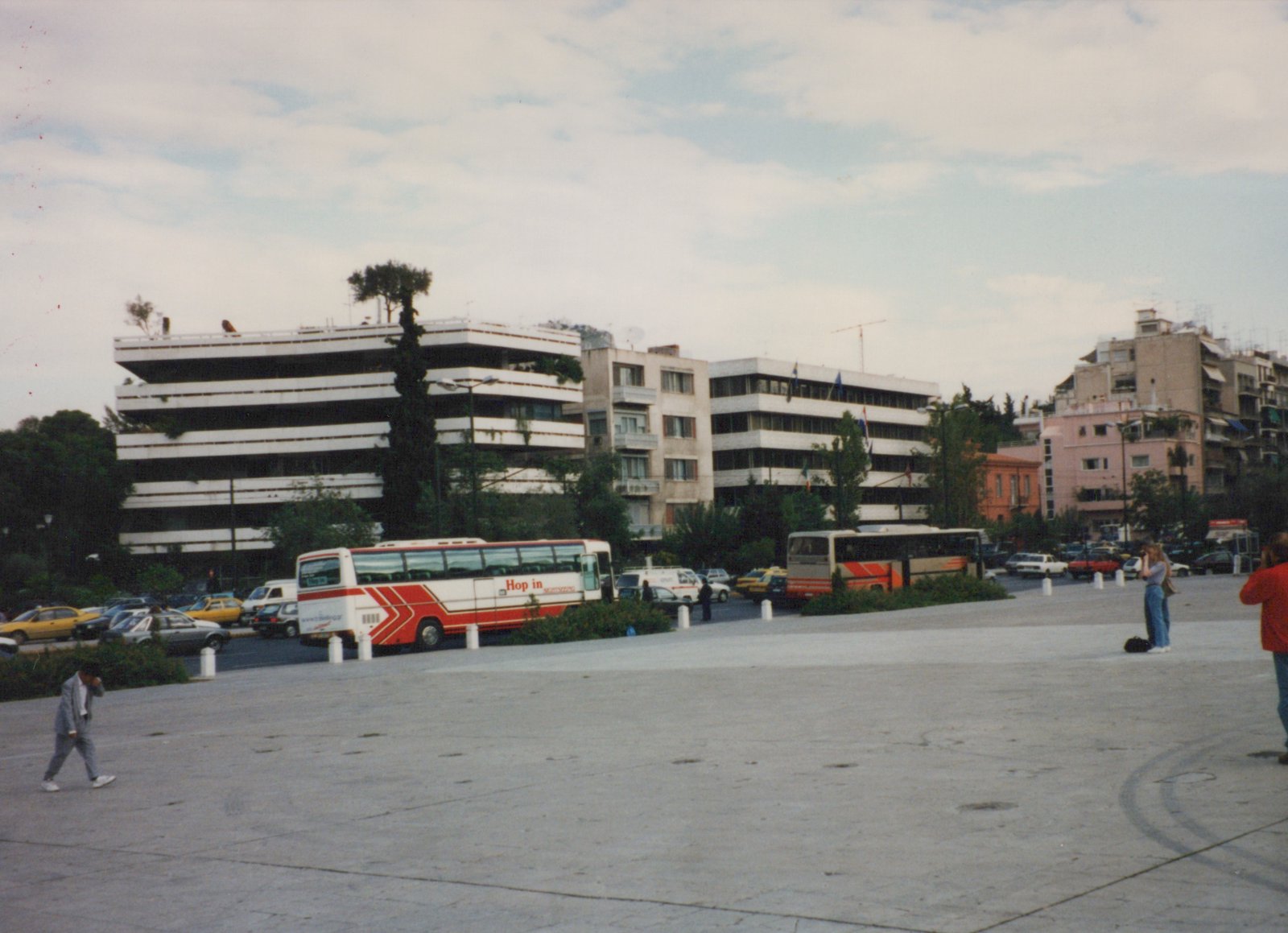 Two tour buses, one white with red stripes and the other beige with orange stripes, both marked 'Hop in', are parked in a city square. Several pedestrians are walking around, and modern buildings line the background under a cloudy sky.