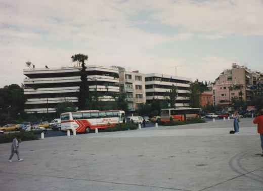 Two tour buses, one white with red stripes and the other beige with orange stripes, both marked 'Hop in', are parked in a city square. Several pedestrians are walking around, and modern buildings line the background under a cloudy sky.