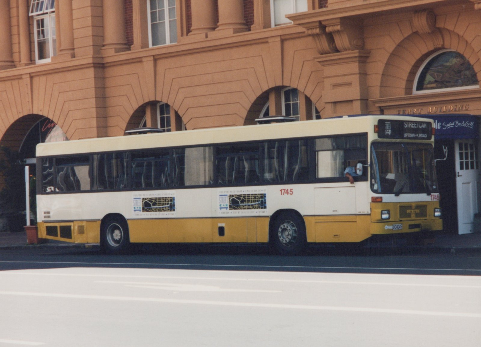 Side view of a white and yellow CTA bus, number 1745, parked on a street in front of a building labeled Ferry Building, with a driver and passenger visible.