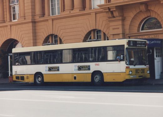 Side view of a white and yellow CTA bus, number 1745, parked on a street in front of a building labeled Ferry Building, with a driver and passenger visible.