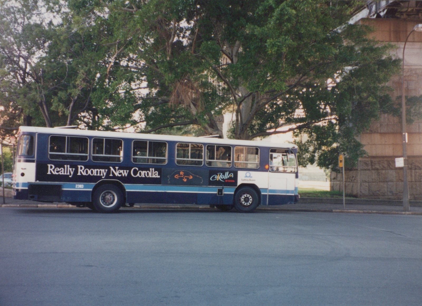 A side view of a blue and white Sydney Buses motor bus, number 2383, with a Toyota Corolla ad, parked beside a road under leafy trees.