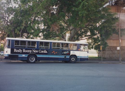 A side view of a blue and white Sydney Buses motor bus, number 2383, with a Toyota Corolla ad, parked beside a road under leafy trees.