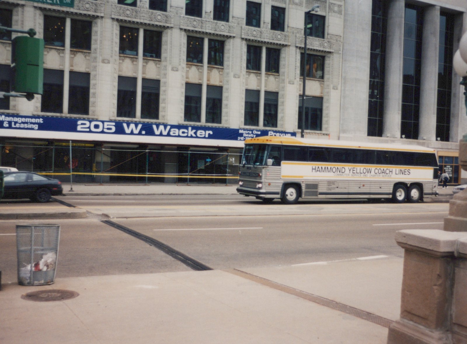 A silver and yellow motor bus with 'Hammond Yellow Coach Lines' lettering on its side is driving down a city street, with a large building behind it bearing the address '205 W. Wacker'.