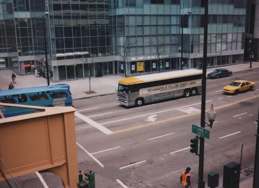 An elevated view of a gray and yellow bus labeled 'Indianapolis Yellow Coach Lines' driving down a city street next to a large glass building, with a green street sign for W Lake St visible.