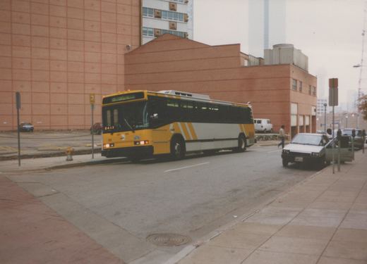 A yellow and white DART bus drives down a city street with brick buildings and parked cars in the background.