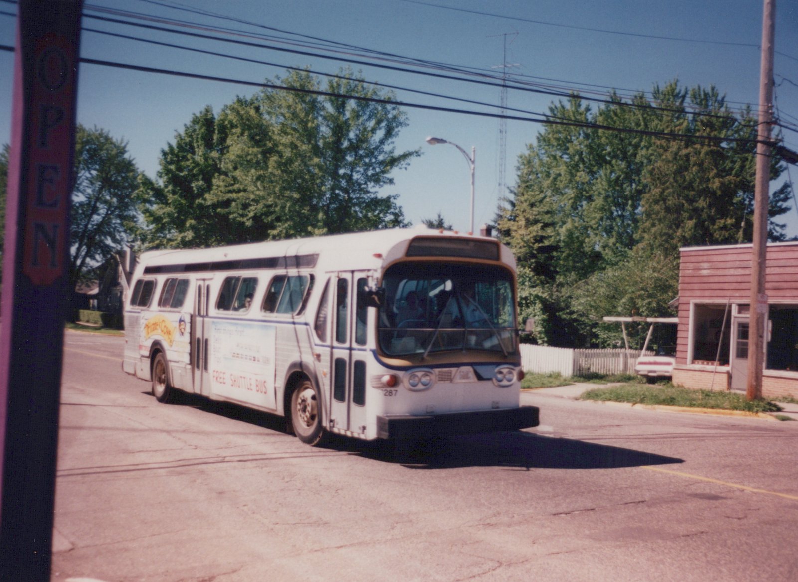 A white bus with 'Pirate's Cove Free Shuttle Bus' lettering and the number 287 on the front drives down a street lined with trees and power lines on a clear, sunny day.