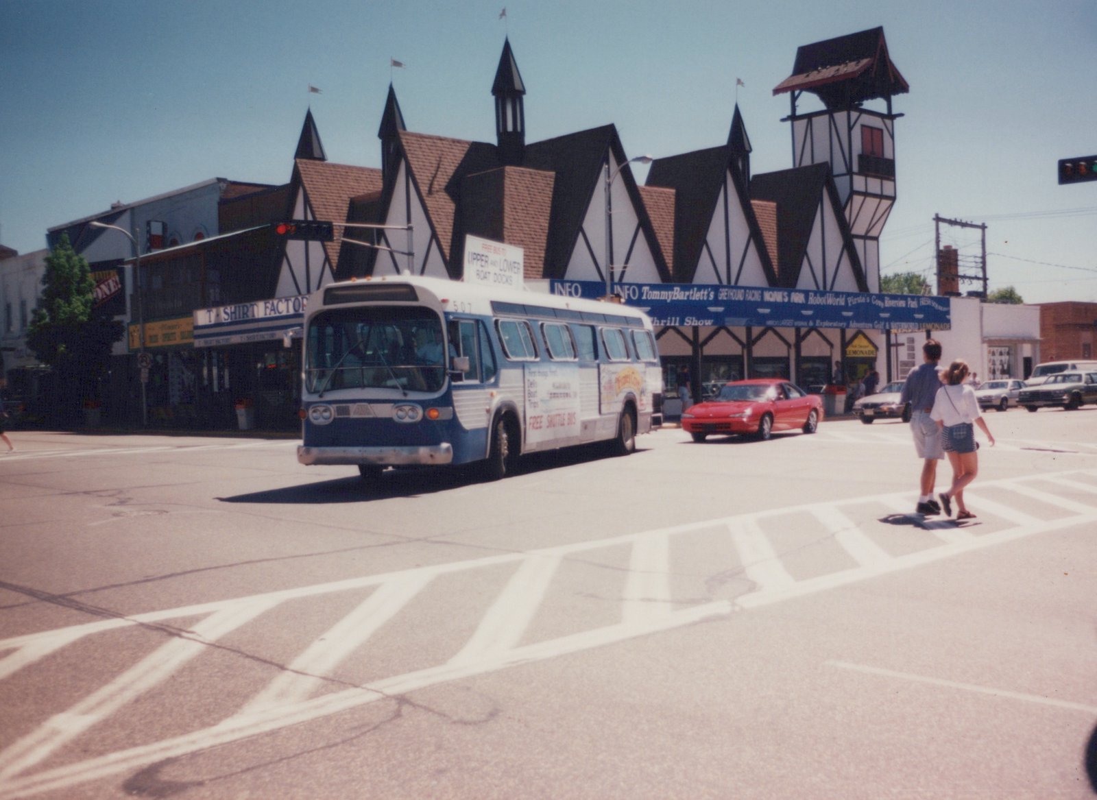 A blue and white shuttle bus, number 507, drives down a street in Wisconsin Dells on a clear, sunny day, with two people crossing the street in front of it.