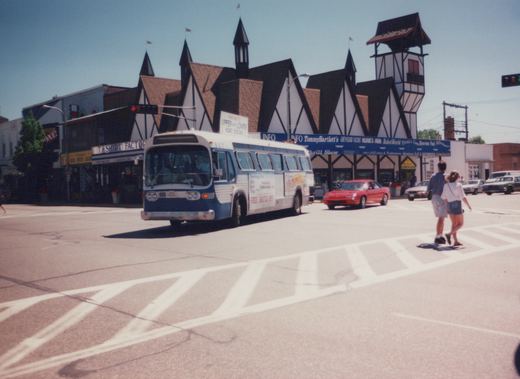 A blue and white shuttle bus, number 507, drives down a street in Wisconsin Dells on a clear, sunny day, with two people crossing the street in front of it.