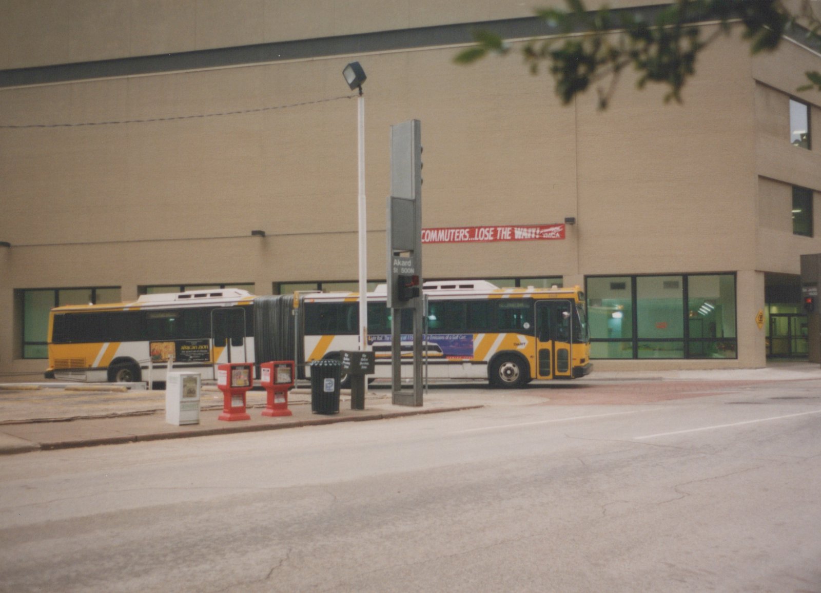 A yellow and white articulated bus with DART lettering is parked on a city street next to a building with a YMCA banner and a street sign for Akard Street.