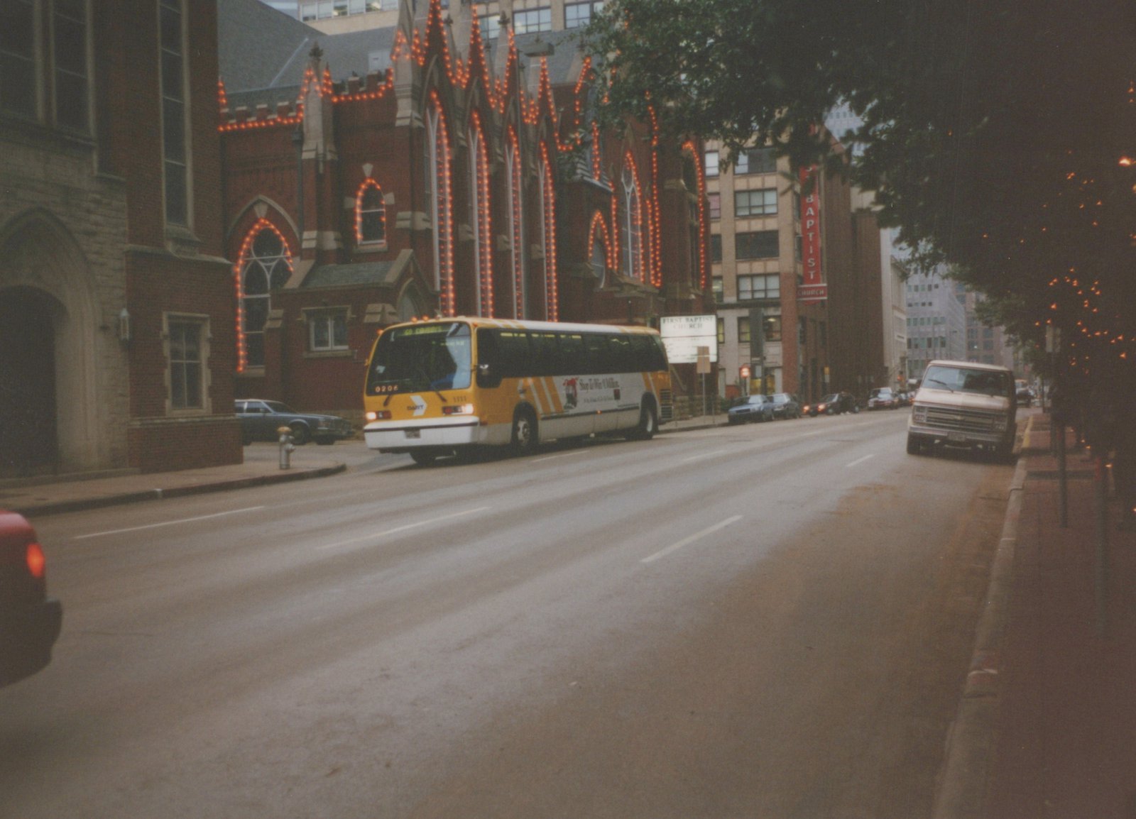 A yellow and white DART bus drives down a wet city street, passing a large brick church with orange string lights and parked cars along the curb.