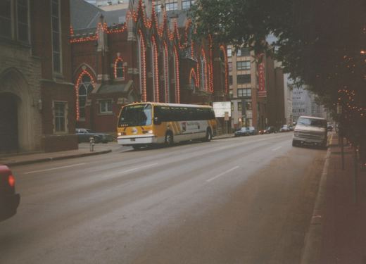 A yellow and white DART bus drives down a wet city street, passing a large brick church with orange string lights and parked cars along the curb.