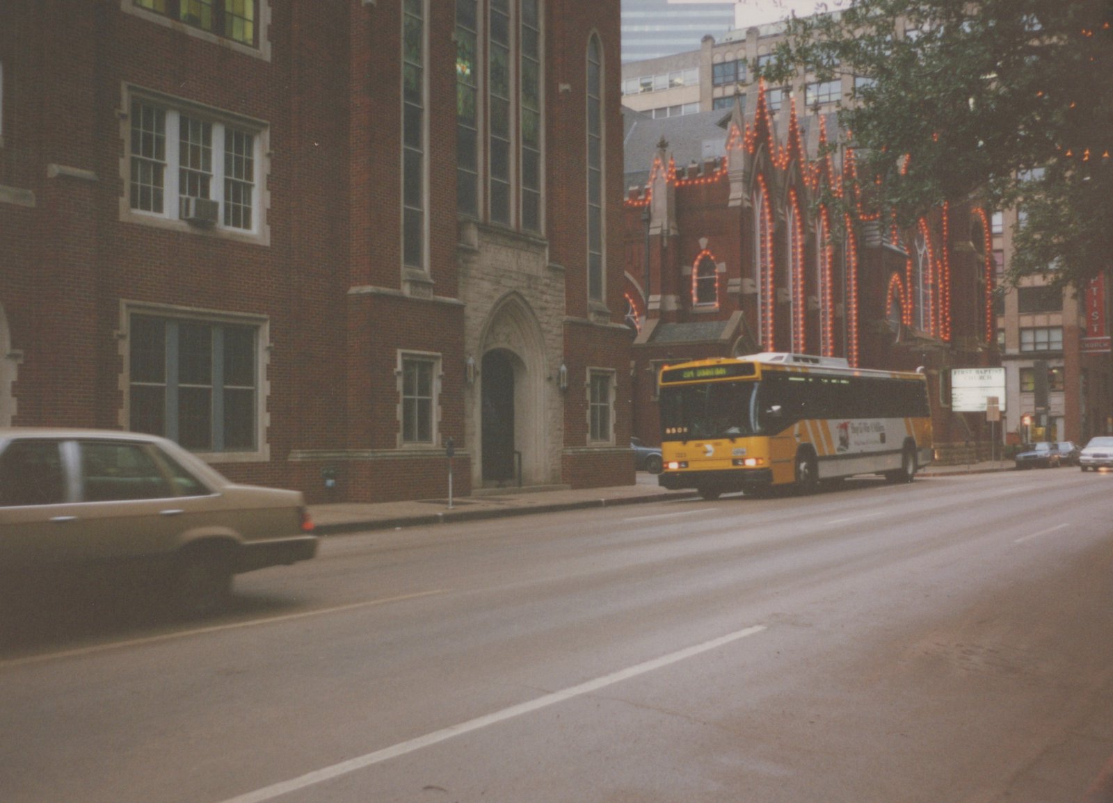 A yellow and white motor bus with the number 3323 on the front is driving down a city street lined with brick buildings. A tan car is blurred in the foreground, and a church with a sign reading 'FIRST BAPTIST CHURCH' is visible in the background.