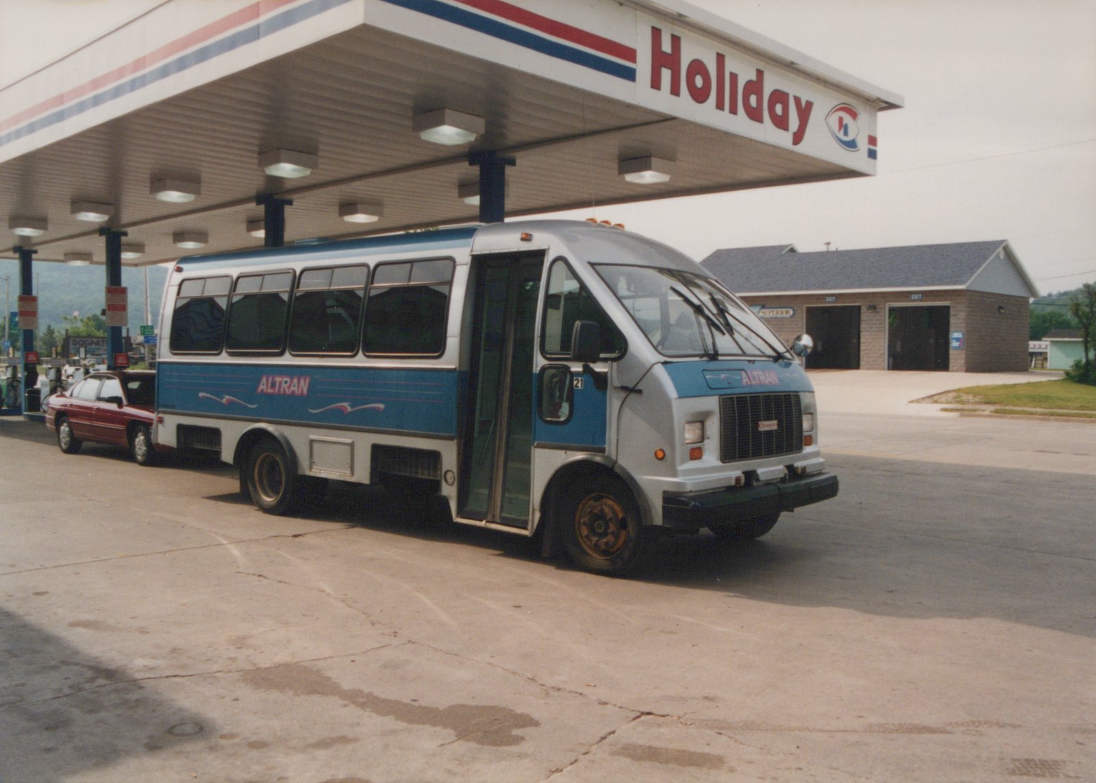 A white and blue ALTRAN motor bus, number 21, is parked under the canopy of a Holiday gas station, with a maroon car visible behind it.