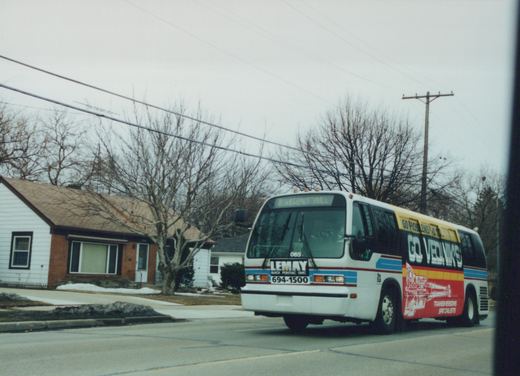 A white Flxible Metro bus with blue and red stripes, marked number 065, drives down a residential winter street with bare trees and patches of snow, its front sign reading '2 KEGENCT MLL'.
