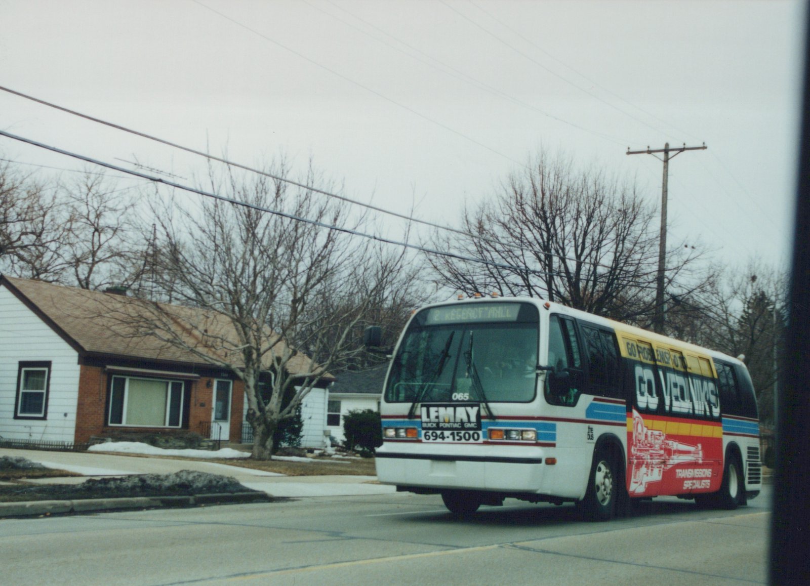 A white Flxible Metro bus with blue and red stripes, marked number 065, drives down a residential winter street with bare trees and patches of snow, its front sign reading '2 KEGENCT MLL'.