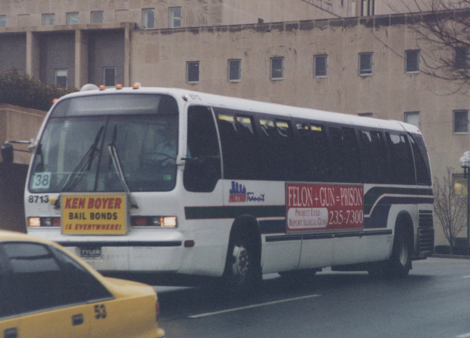 A white city bus with the number 8713 and 'METRO Transit' on its side, driving down a street with a yellow taxicab in the foreground and a large building in the background.