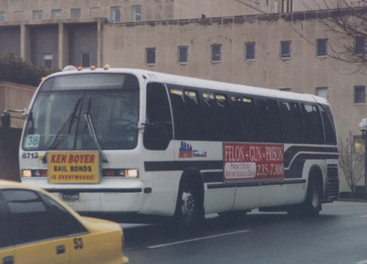 A white city bus with the number 8713 and 'METRO Transit' on its side, driving down a street with a yellow taxicab in the foreground and a large building in the background.
