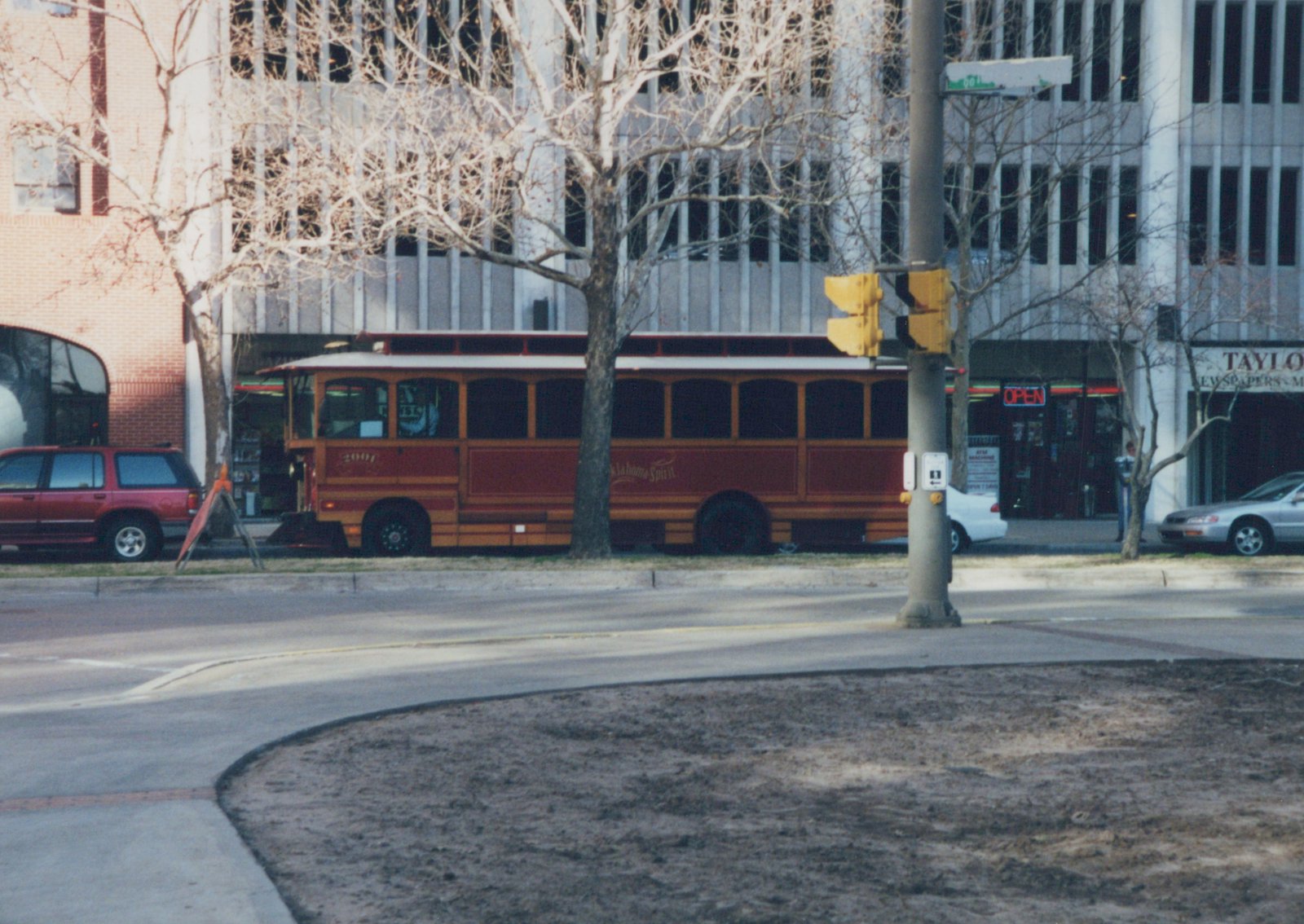 A red and gold trolley-style bus with the fleet number 2001 and the words 'Oklahoma Spirit' on its side is parked on a city street lined with bare trees and modern buildings.