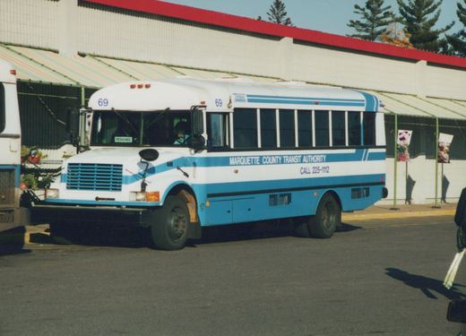 A side view of a white and blue Marquette County Transit Authority bus with the number 69 on the front, parked outdoors on a sunny day.