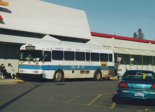 A white and blue Flxible Metro bus, number 109, for the Marquette County Transit Authority is parked at a terminal with people nearby and a blue car in the foreground.