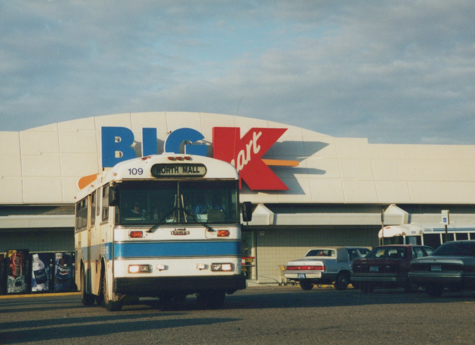 A white and blue Flxible Metro bus with the number 109 and a destination sign reading 'NORTH/MALL' is parked on a street in front of a Big K store. A second bus and several cars are visible in the background.