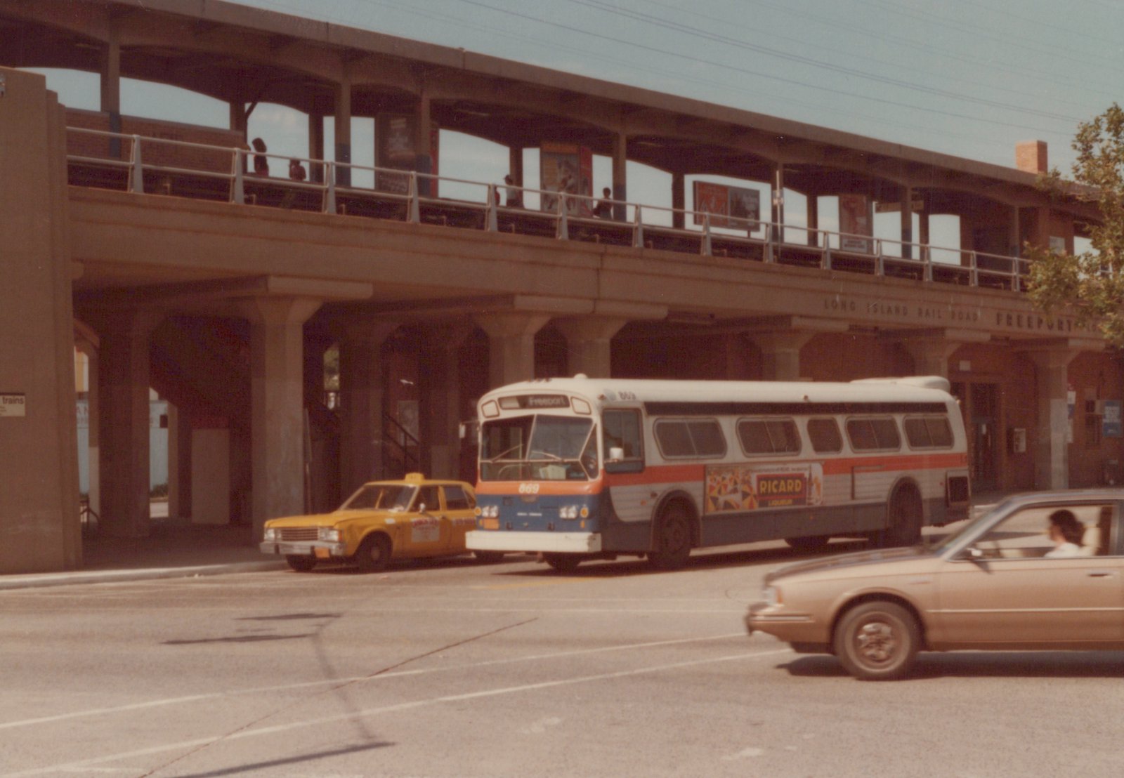 A white, orange, and blue bus with the number 869 and 'Freeport' on its sign is parked at a street-level station. Behind it, a yellow taxi and a tan car are visible. Above, people stand on an elevated train platform marked 'LONG ISLAND RAIL ROAD FREEPORT'.