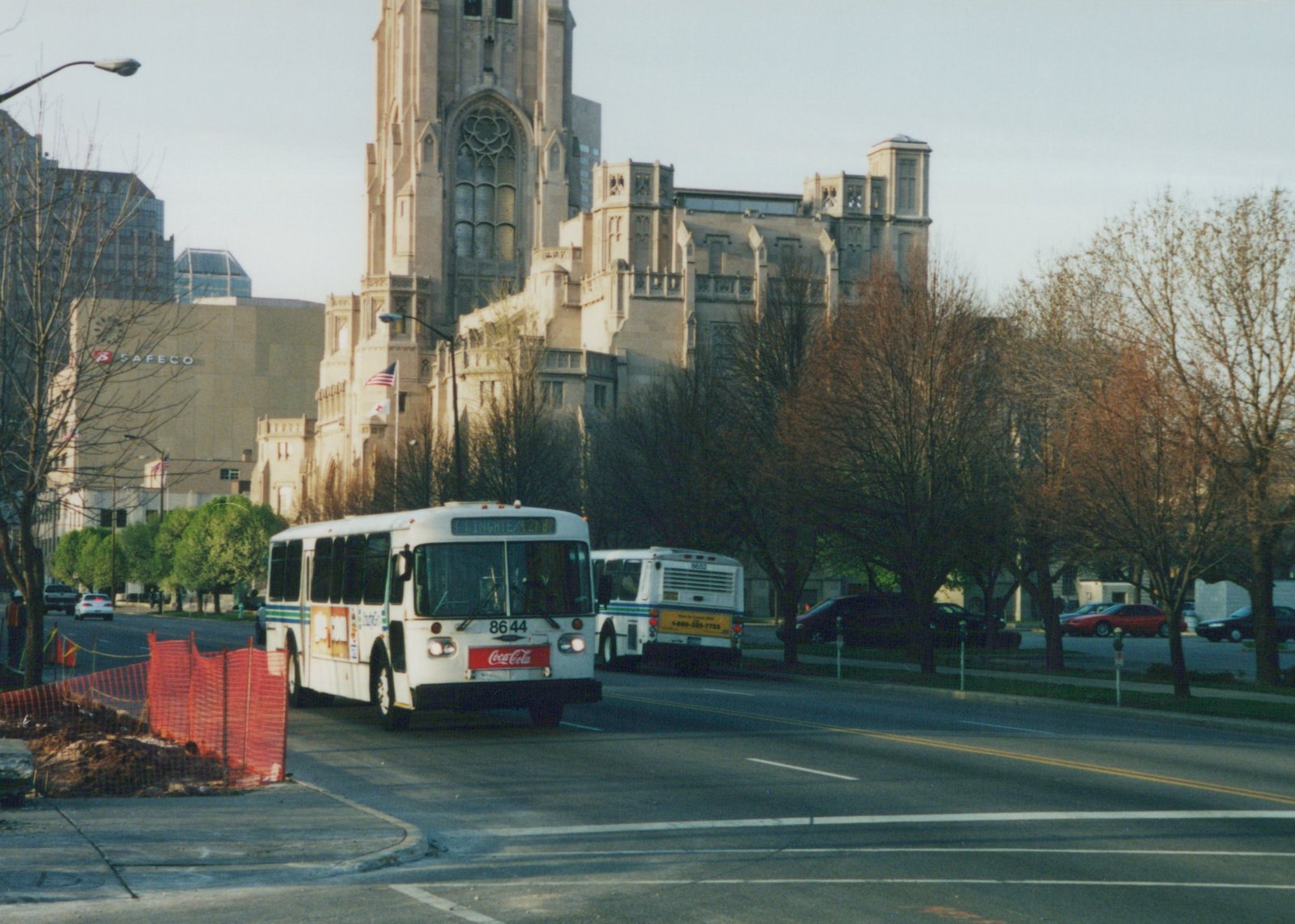 A white Indianapolis bus with green and blue stripes, numbered 8644, drives down a city street with trees and a large stone church in the background.