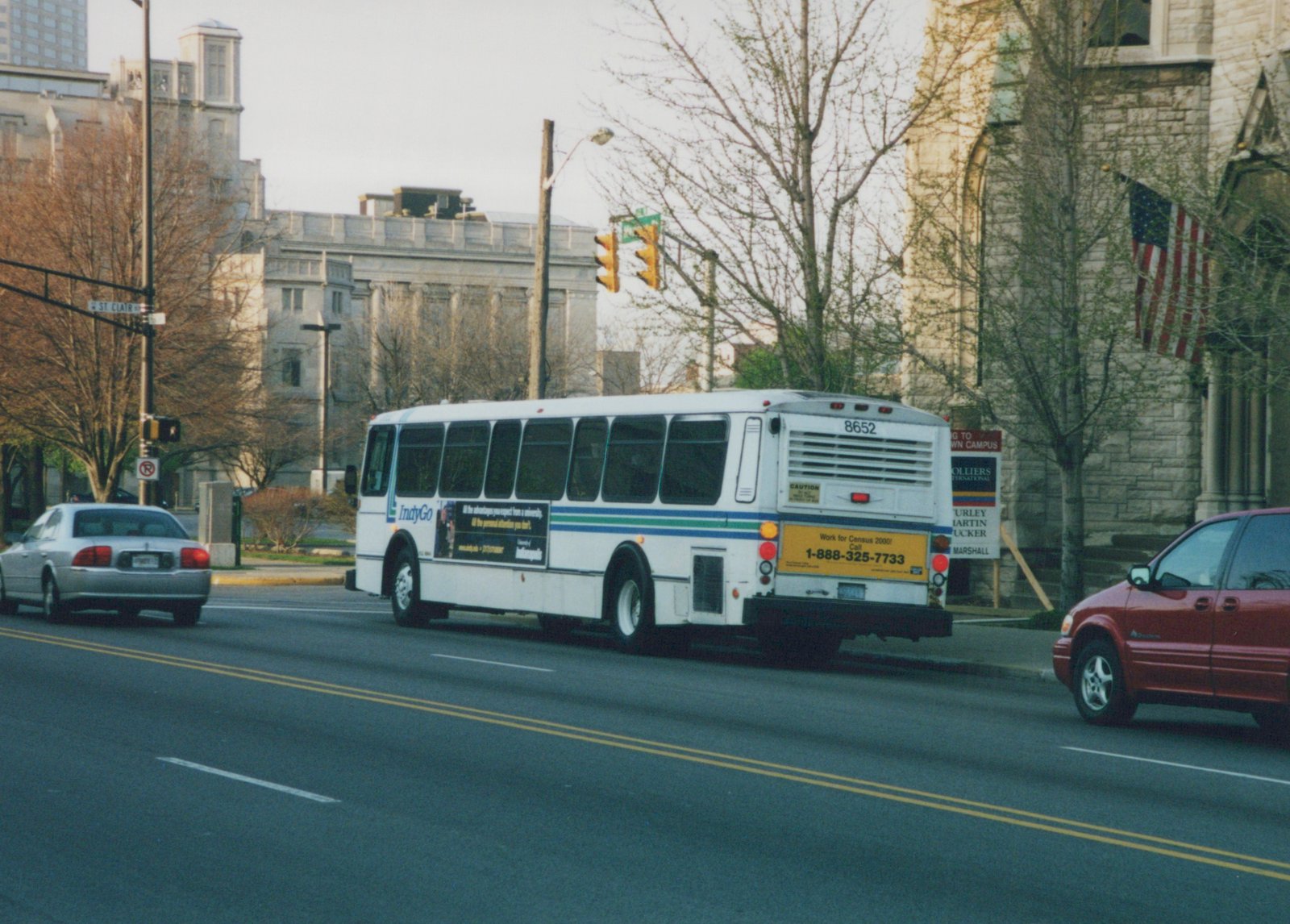 A white IndyGo bus with fleet number 8652, carrying a 'Census 2000' ad, travels down a city street lined with trees and buildings.