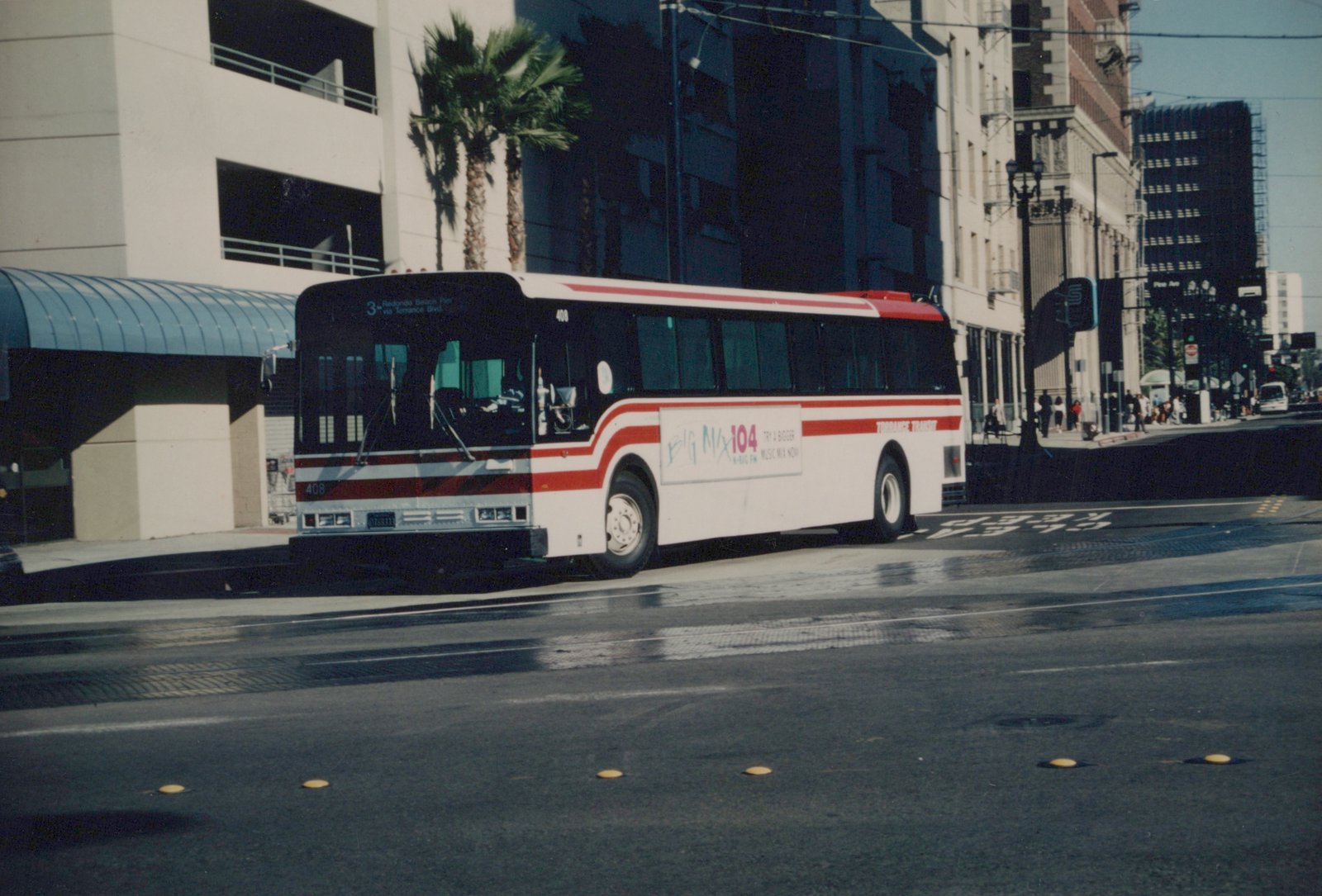 A white and red Torrance Transit bus, number 408, is shown driving on a city street with palm trees and buildings in the background. The destination sign reads '3 Redondo Beach Pier via Torrance Blvd'.