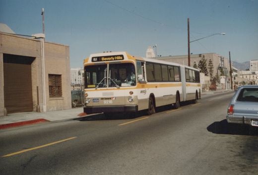 A white and orange RTD articulated bus, numbered 9216, drives down a city street under a clear blue sky, with a light blue car visible beside it.